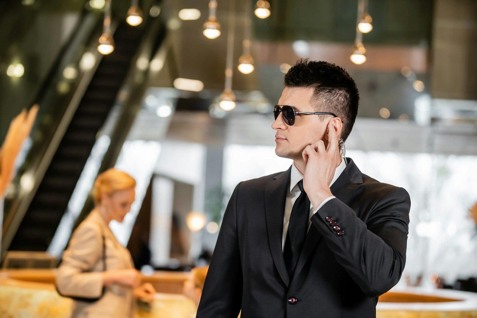 bodyguard in sunglasses, handsome man in suit and tie touching earpiece in lobby of hotel